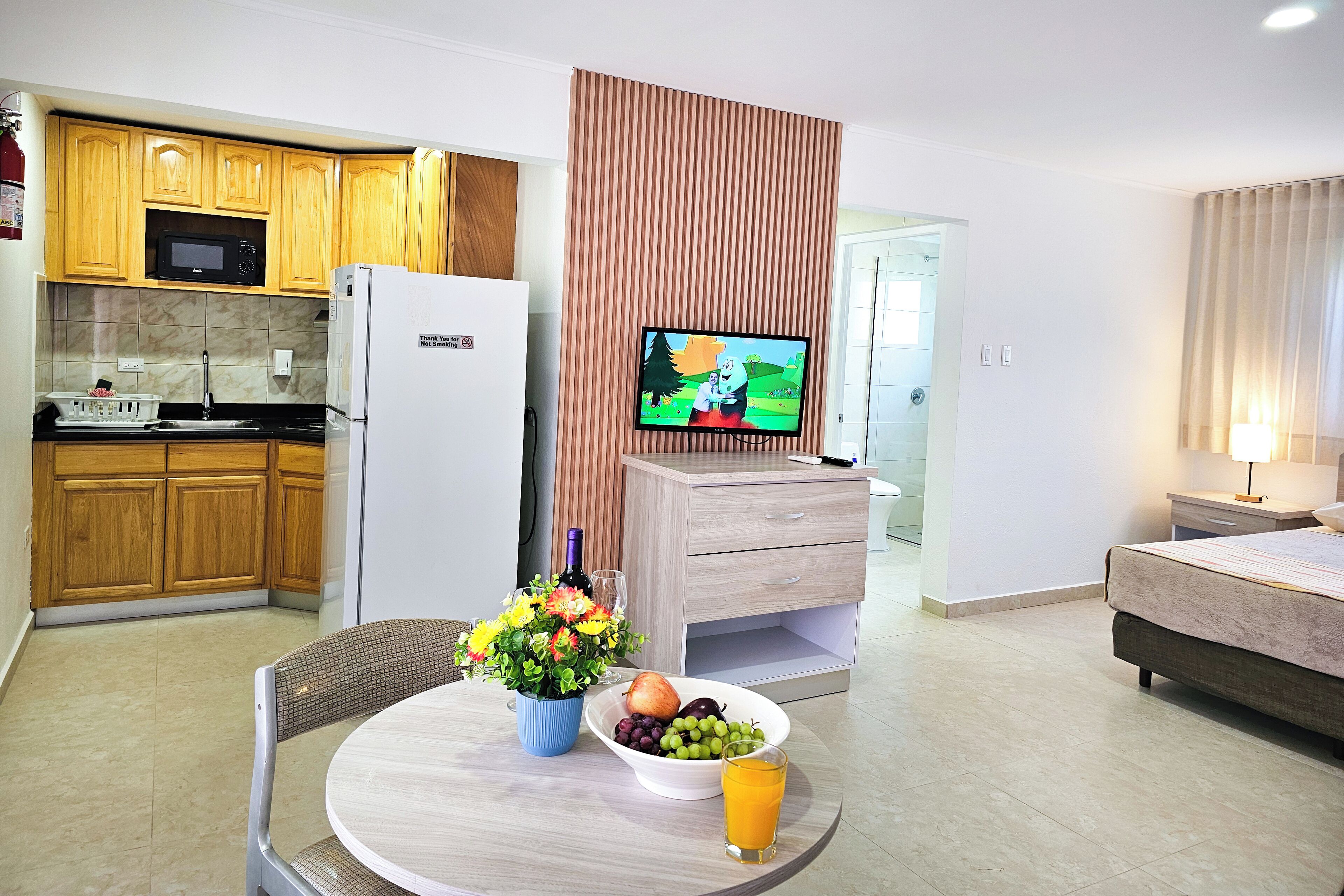 Une cuisine moderne avec des meubles en bois, un réfrigérateur blanc et un four à micro-ondes. On y trouve un coin repas avec une table, des chaises et un vase de fleurs. Un téléviseur est fixé sur un panneau en bois, et on aperçoit un lit à l’arrière-plan.