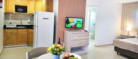 Cocina moderna con armarios de madera, un refrigerador blanco y un microondas. Hay un comedor con una mesa, sillas y un florero. Un televisor está montado en un panel de madera, y se ve una cama al fondo.
