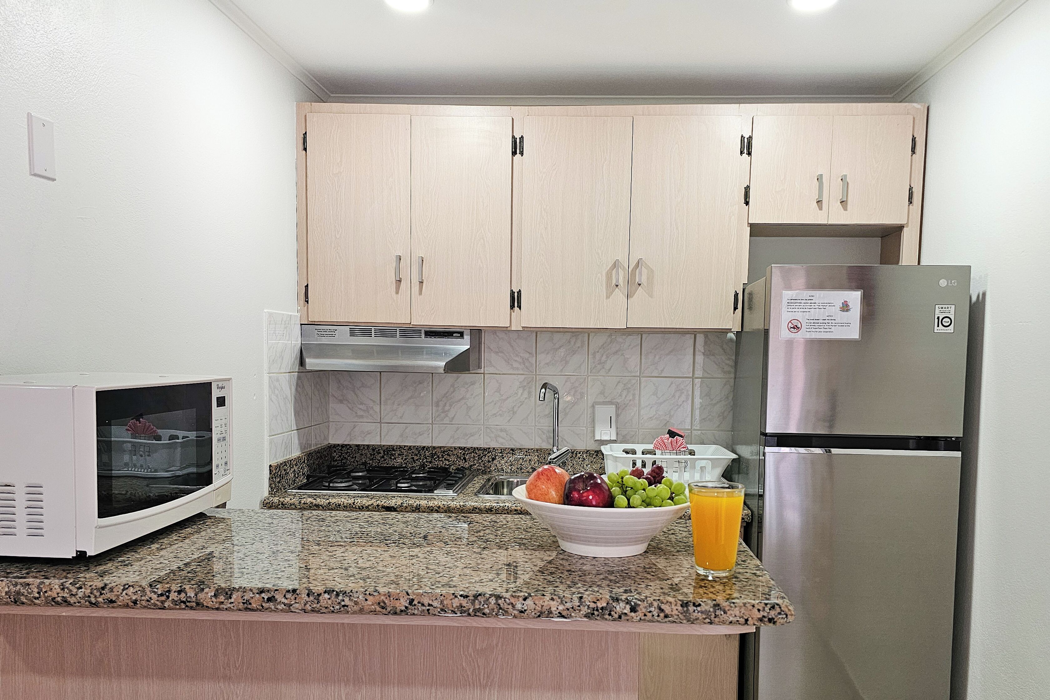 Une cuisine avec des meubles de couleur bois clair, un réfrigérateur en acier inoxydable, un four à micro-ondes, une cuisinière à gaz et un saladier rempli de fruits et de jus.