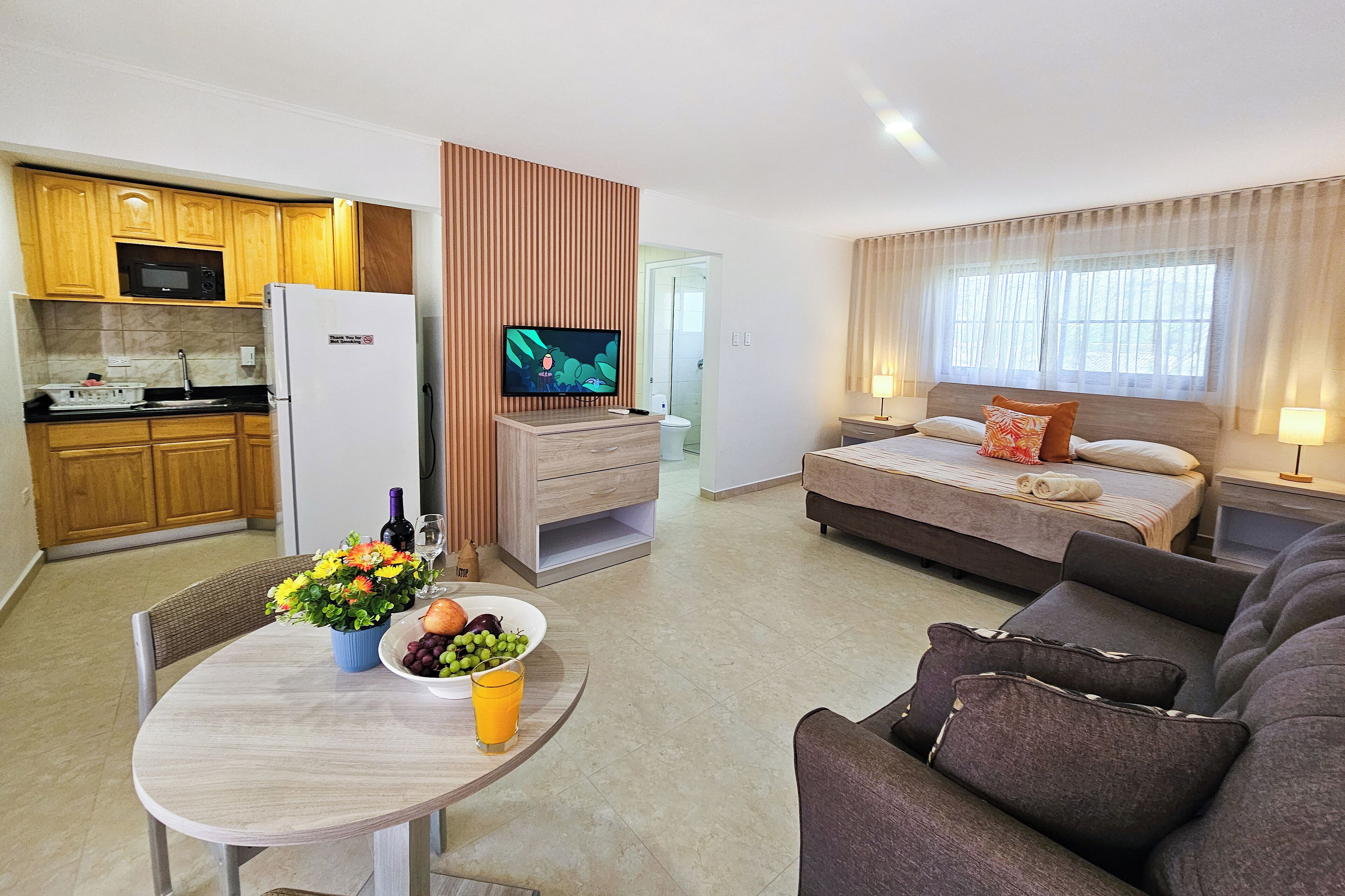 Una habitación de hotel moderna con kitchenette, un televisor de pantalla plana, una cama con mesita de noche y una zona de comedor con una mesa preparada para dos.