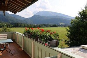 A patio with a table and chairs, a view of green fields and mountains, and a wooden pergola.