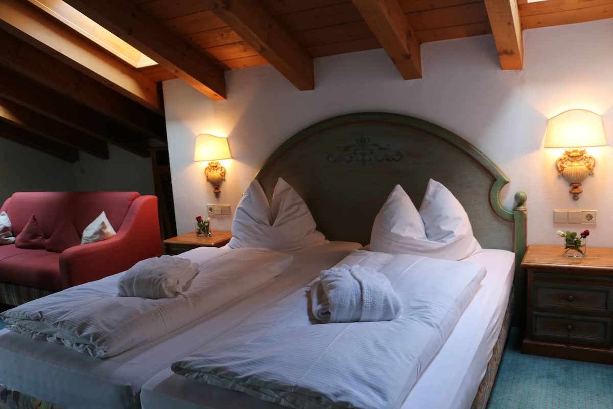 A double bed with white linens, two bedside tables with lamps, a red armchair, and wooden ceiling beams.