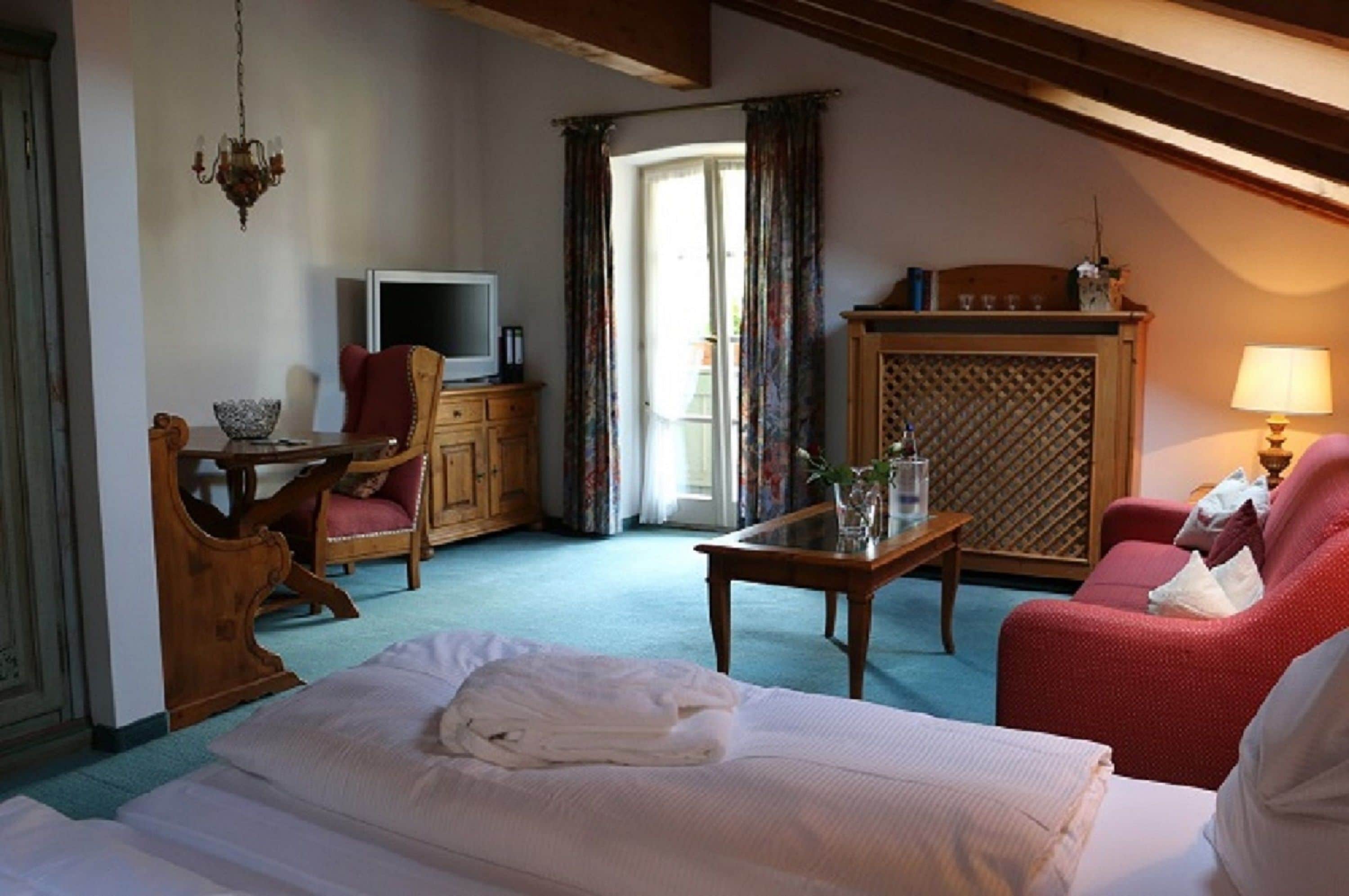 A hotel room with a bed, a desk with a television, a red armchair, a wooden cabinet, and a window with curtains.