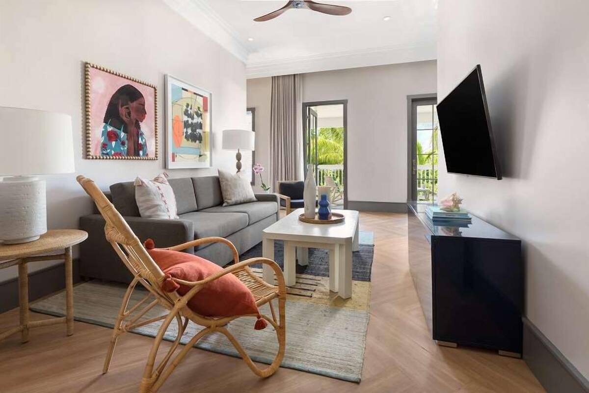 A modern living room with a grey sofa, a wicker chair, a white coffee table, and a flat-screen TV mounted on the wall.