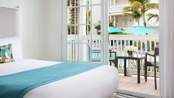 A hotel room with a balcony overlooking a pool area, featuring a bed, a table with chairs, and a view of the pool and lounge chairs.