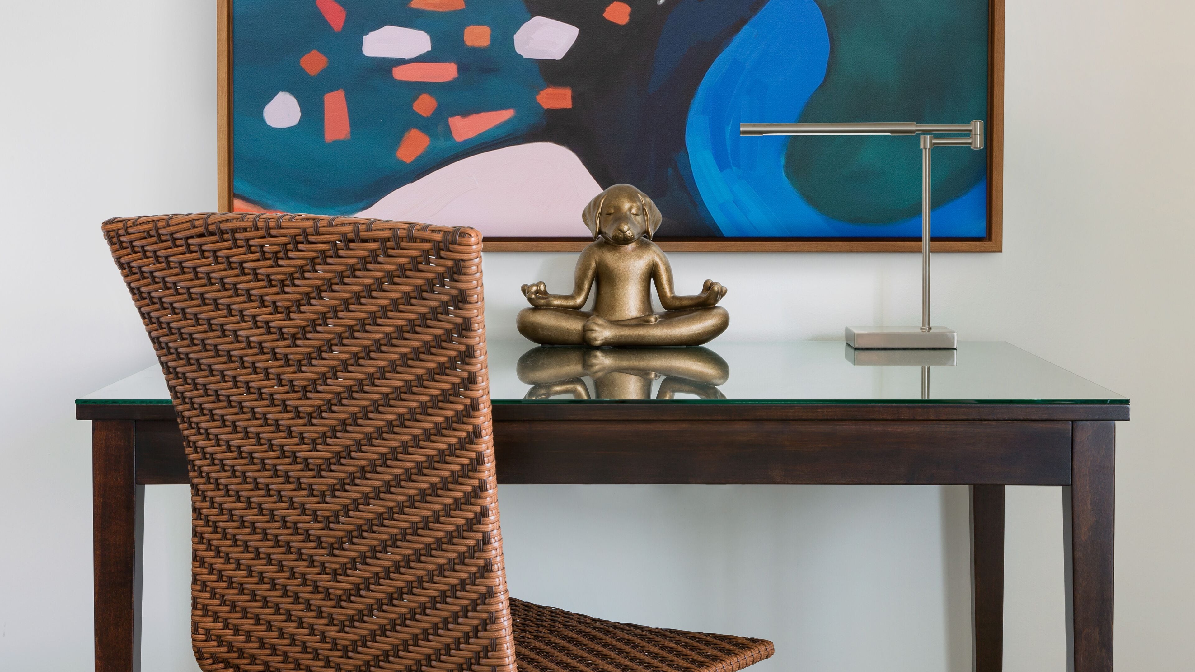 Una silla de mimbre con una estatua de meditación sobre una mesa de vidrio frente a una pared con arte abstracto.