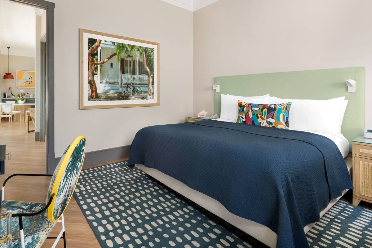 A hotel room with a bed, a desk, a chair, a patterned rug, and a painting on the wall.