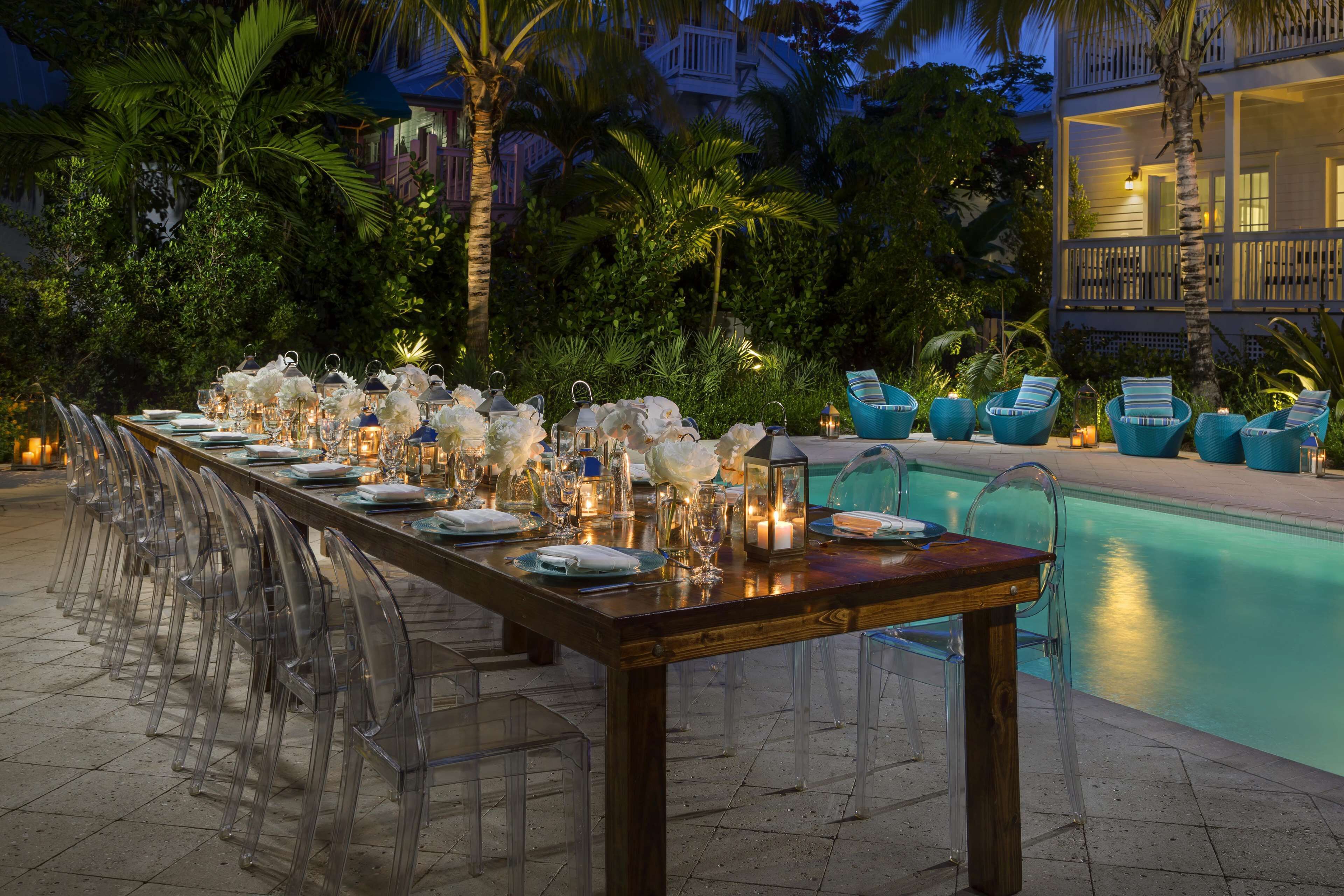 Una larga mesa de comedor preparada para un evento formal, con sillas traslúcidas, velas y cubertería.