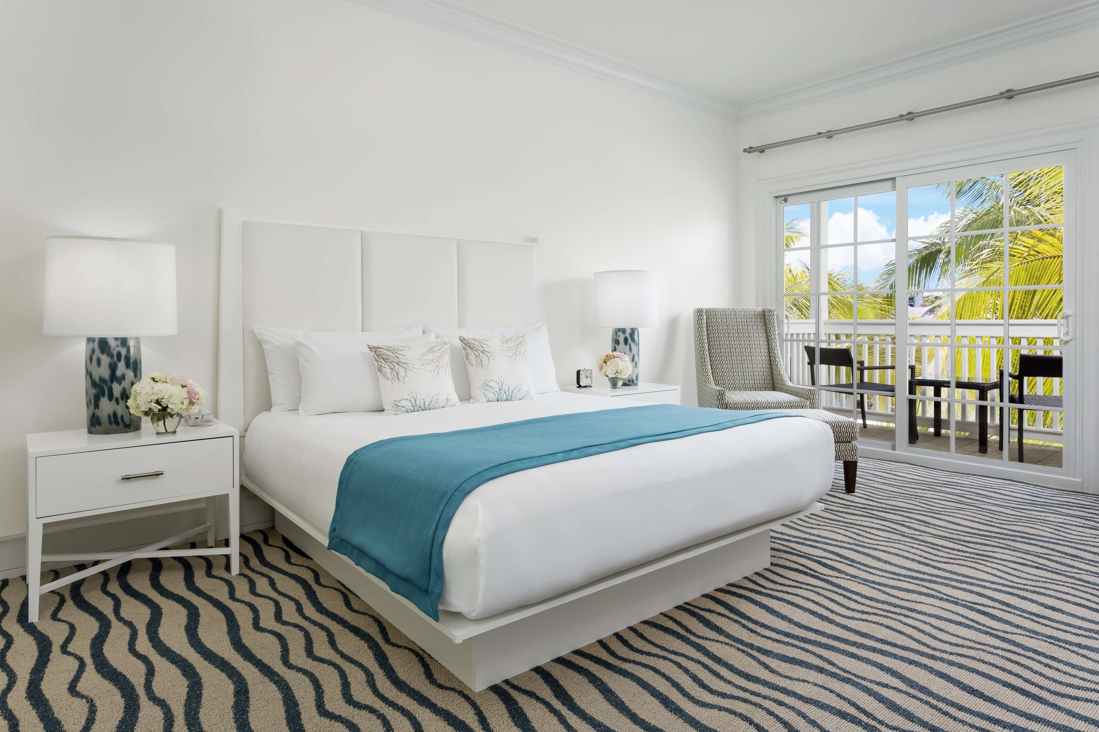 A bedroom with a large bed, white nightstands, a chair, and a balcony with a view of palm trees.