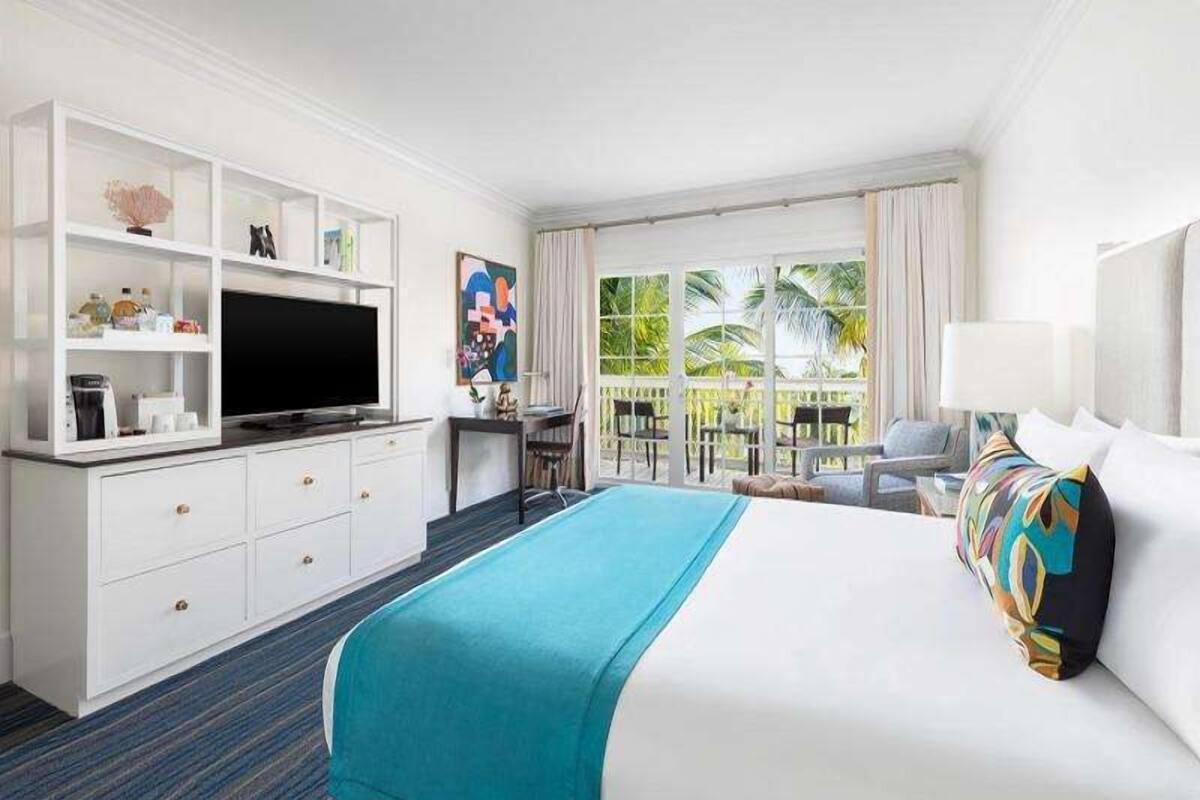 A hotel room with a bed, a desk, a TV, a balcony with a view of palm trees, and a window with curtains.