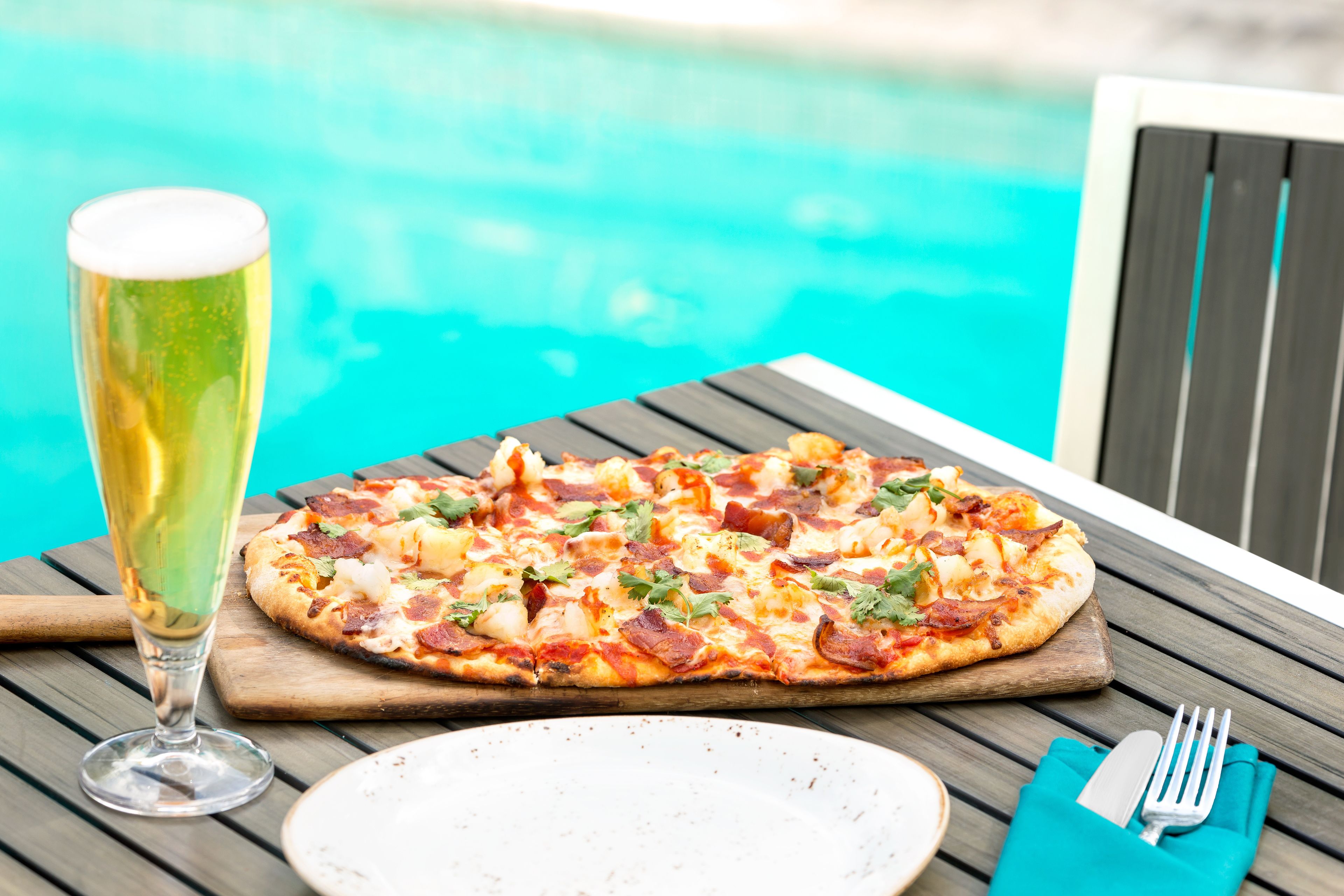 Une pizza servie sur une planche en bois, accompagnée d’un verre de bière, près d’une piscine.