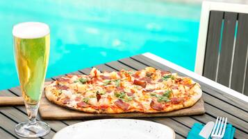 Una pizza sobre una tabla de madera con un vaso de cerveza a su lado, cerca de una piscina.