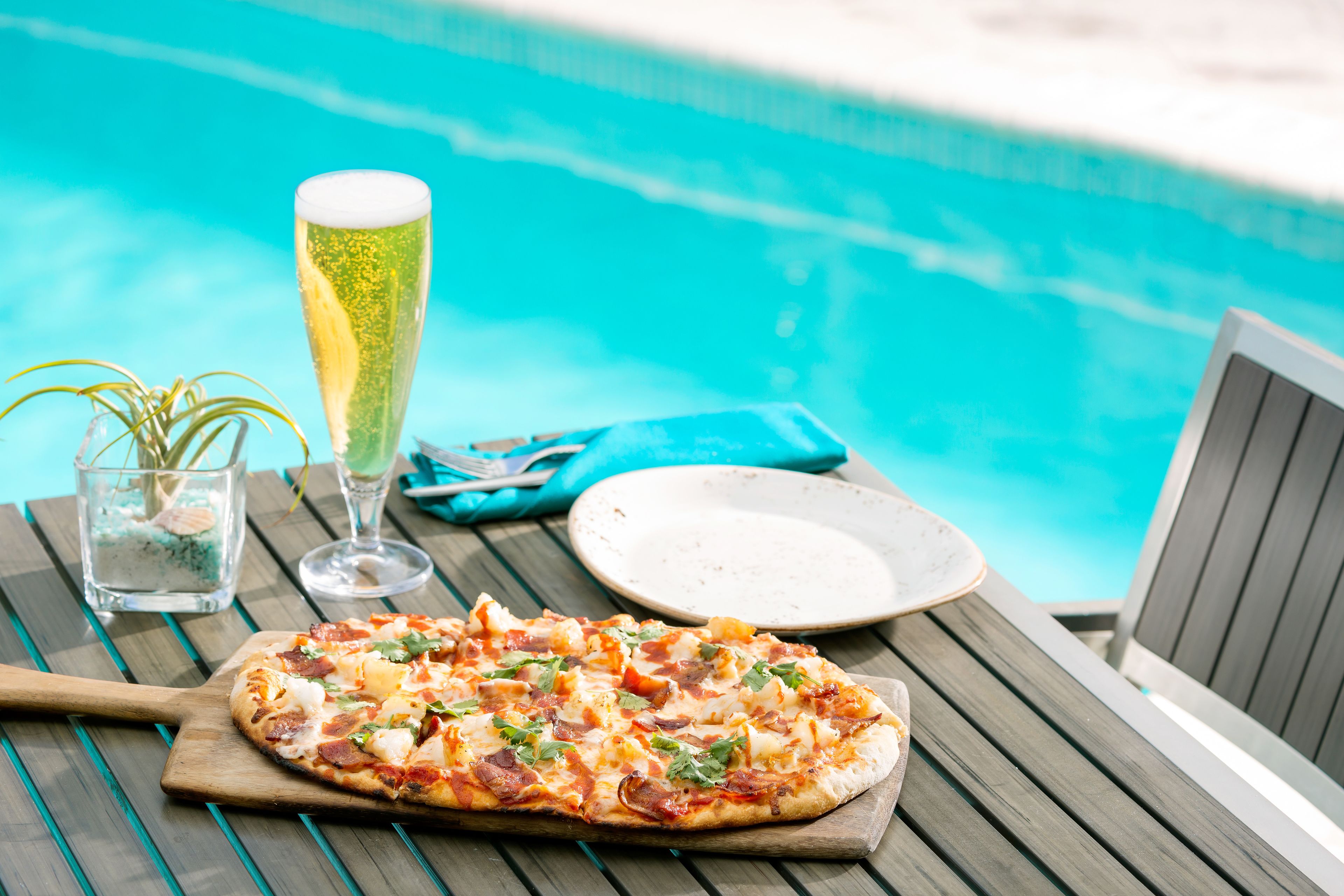 A wooden table with a pizza, a glass of beer, and a plate by a poolside.