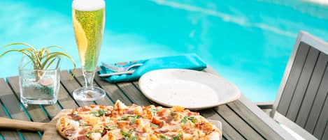 A wooden table with a pizza, a glass of beer, and a plate by a poolside.