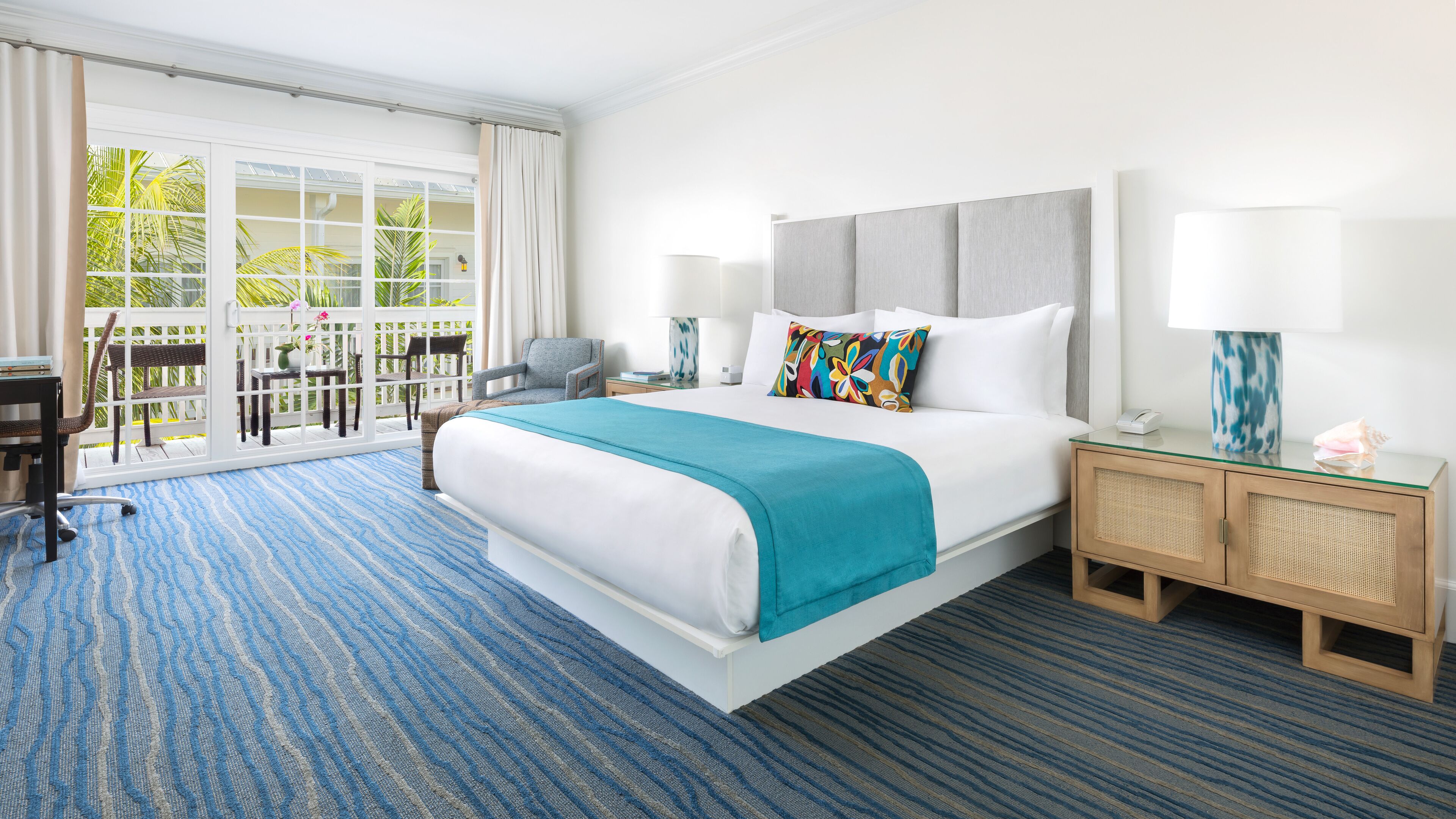 A hotel room with a large bed, a desk, a chair, a nightstand, and a view of a balcony with plants.