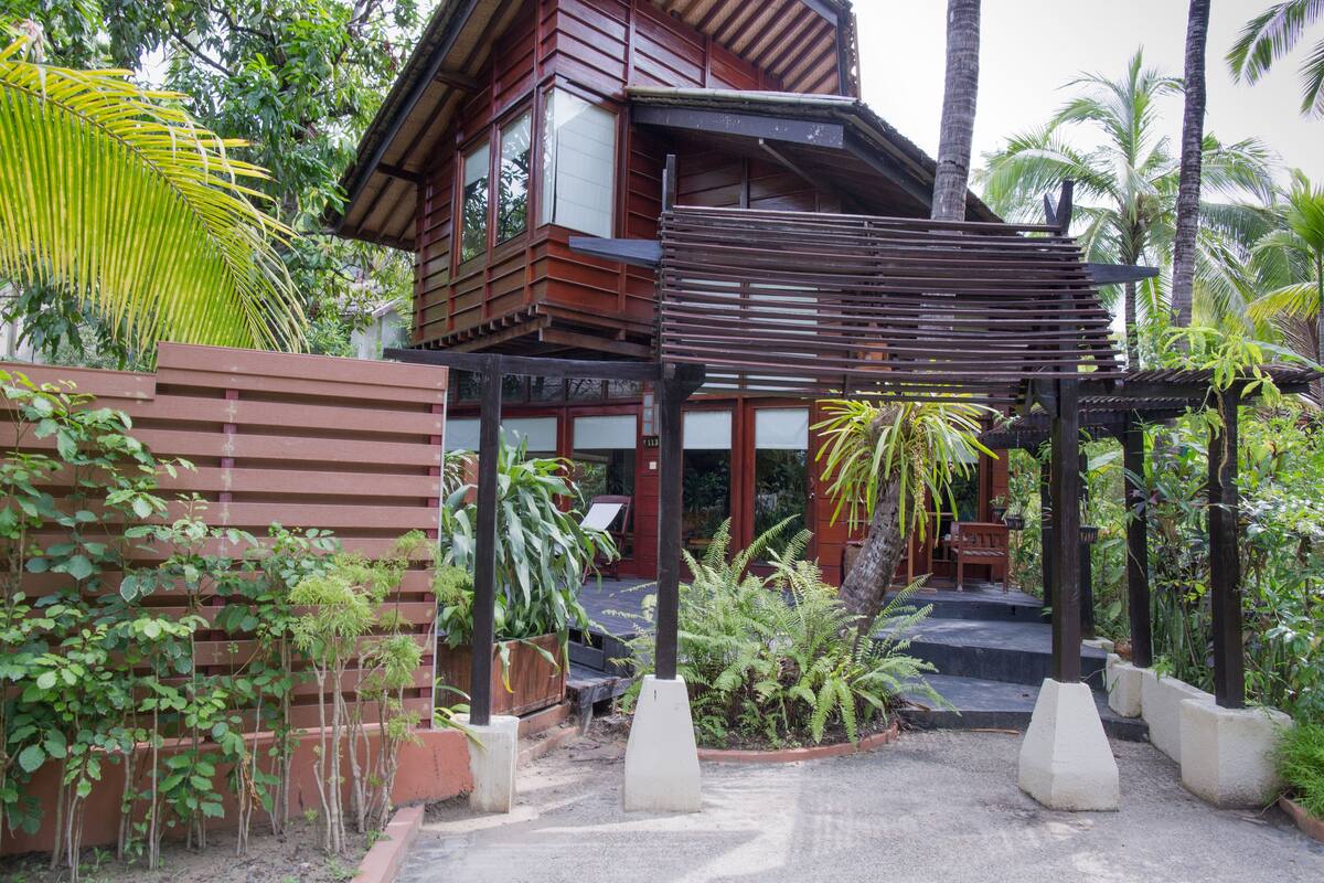 A traditional wooden house with a balcony, surrounded by lush greenery and palm trees.