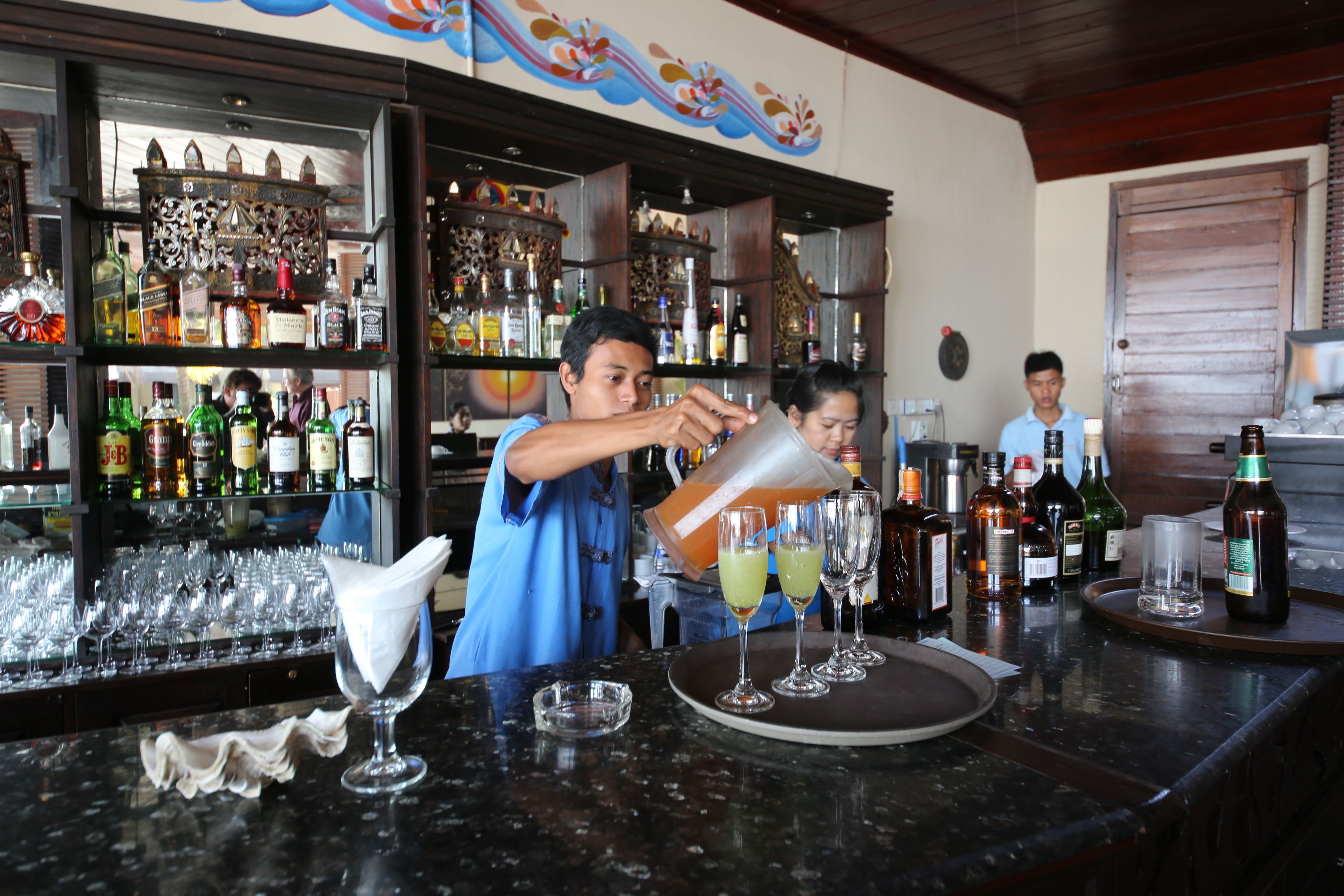 Una zona de bar con estanterías llenas de botellas y vasos, un camarero preparando bebidas y una pared decorativa.