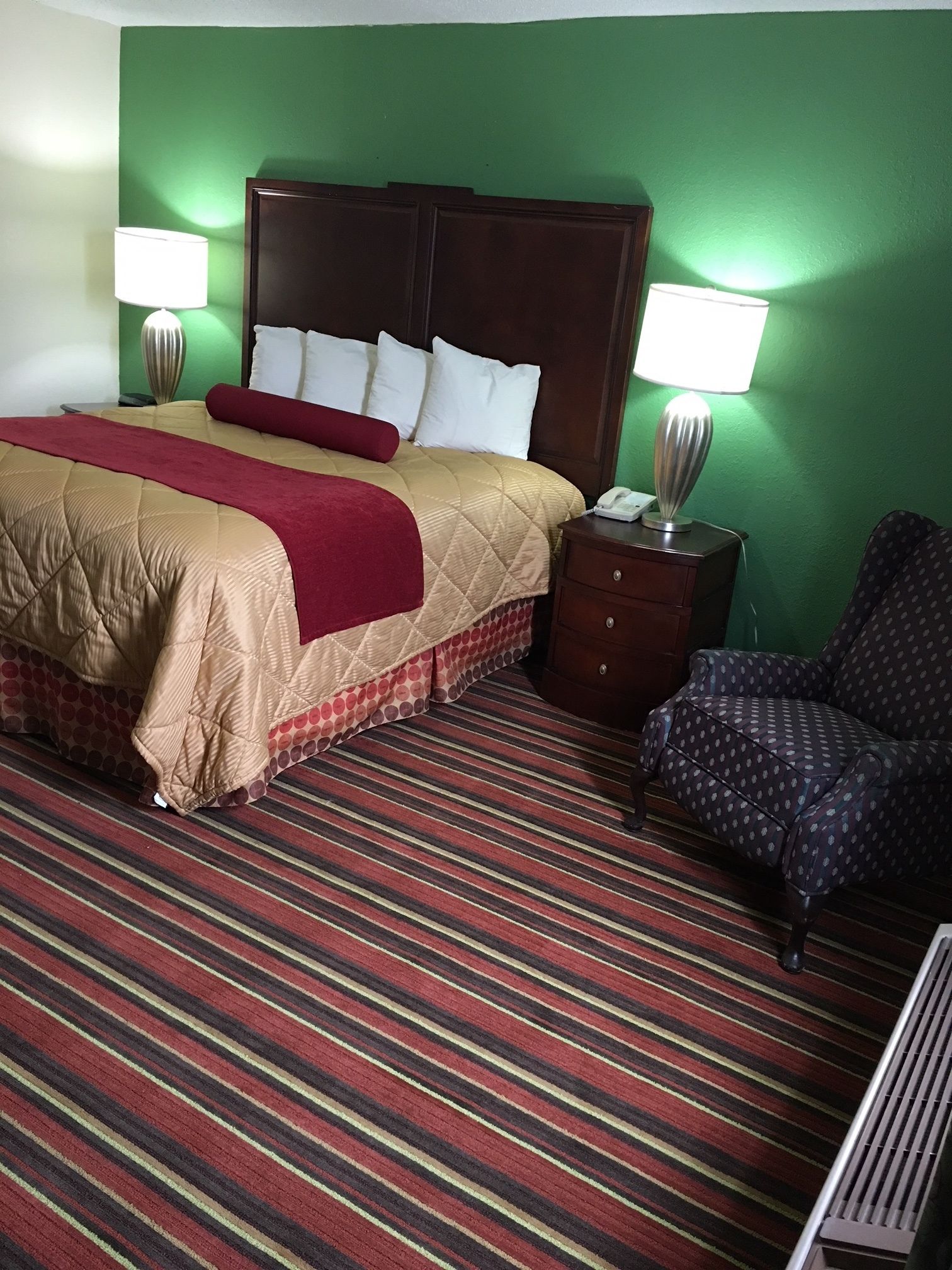 A hotel room with a bed, a nightstand, a chair, and a striped carpet.