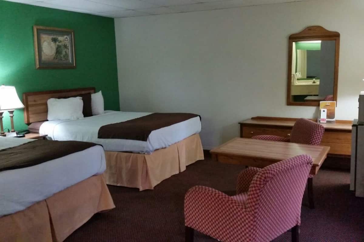 Quarto de hotel com duas camas, uma escrivaninha, uma cadeira, um espelho e um abajur.