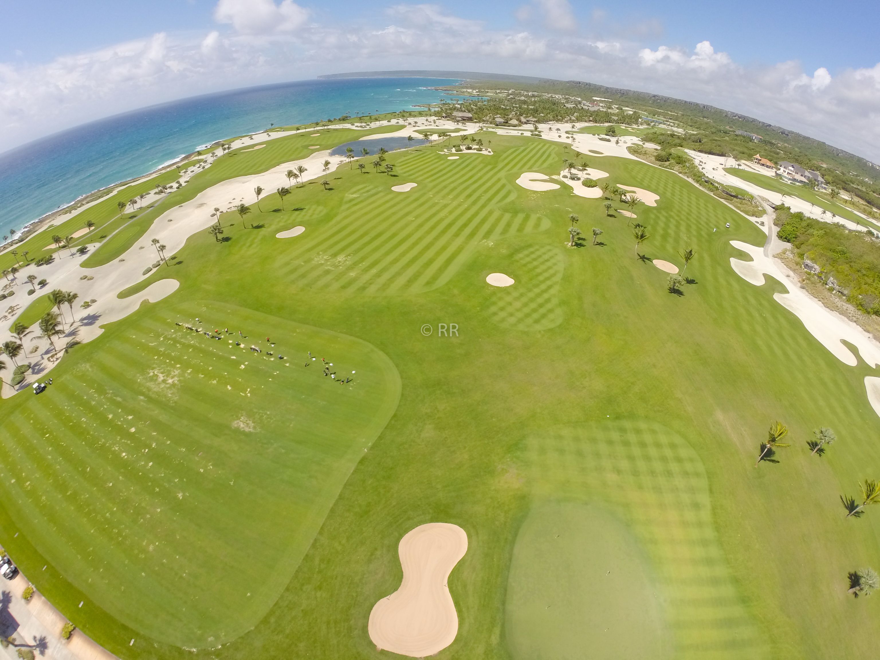 An aerial view of a golf course with sand bunkers, a water hazard, and a view of the ocean and hills in the background.
