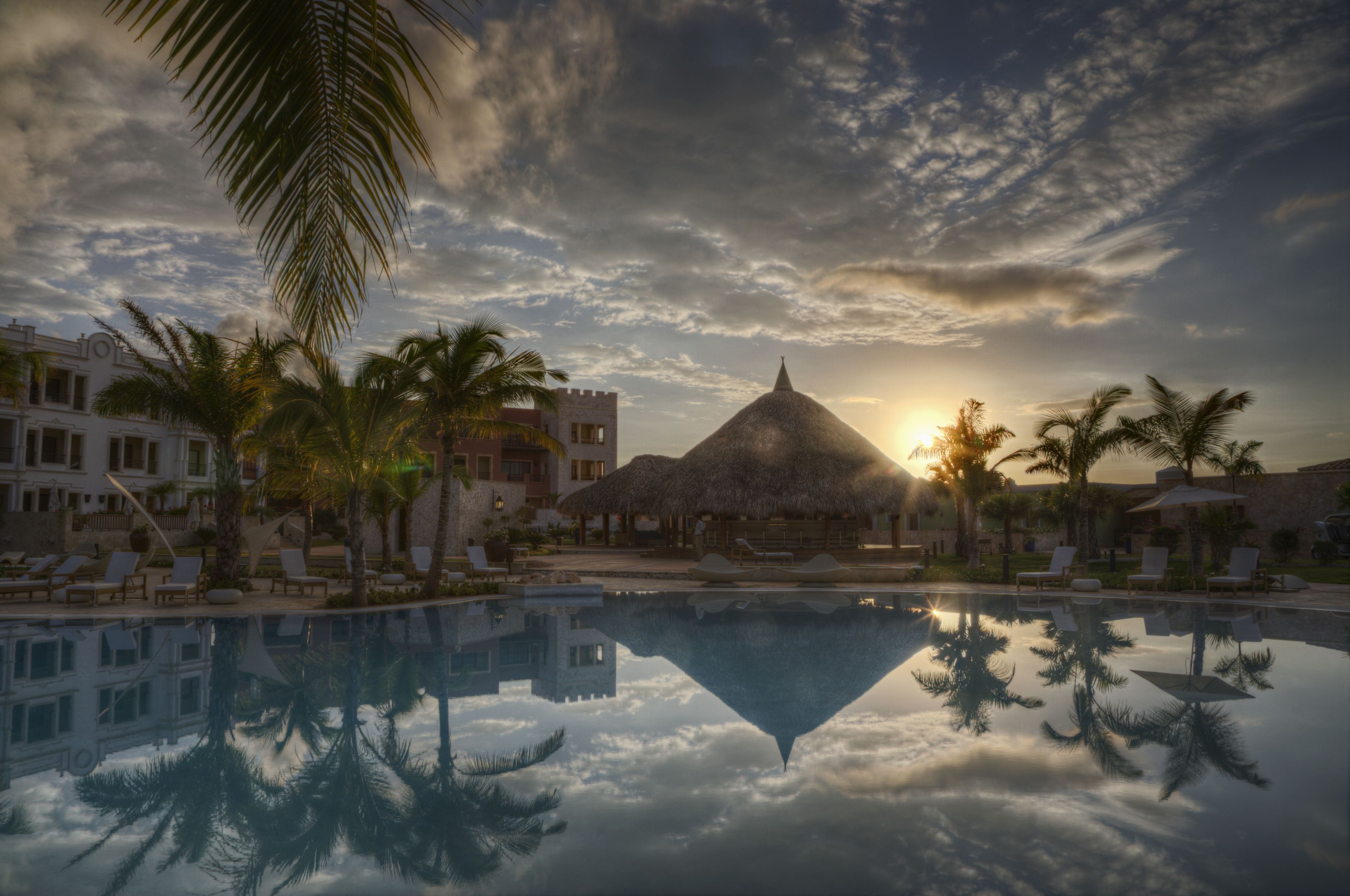 Ein Resort mit Pool, der Palmen und ein Gebäude mit Reetdach spiegelt.