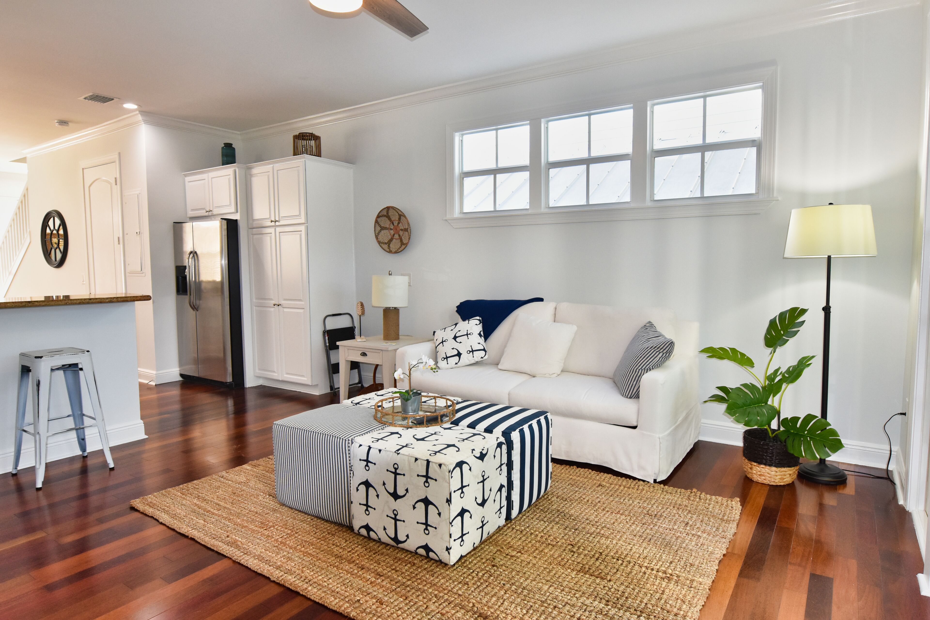 A modern living room with a white sofa, striped ottoman, and a dining area with white cabinets and a refrigerator.