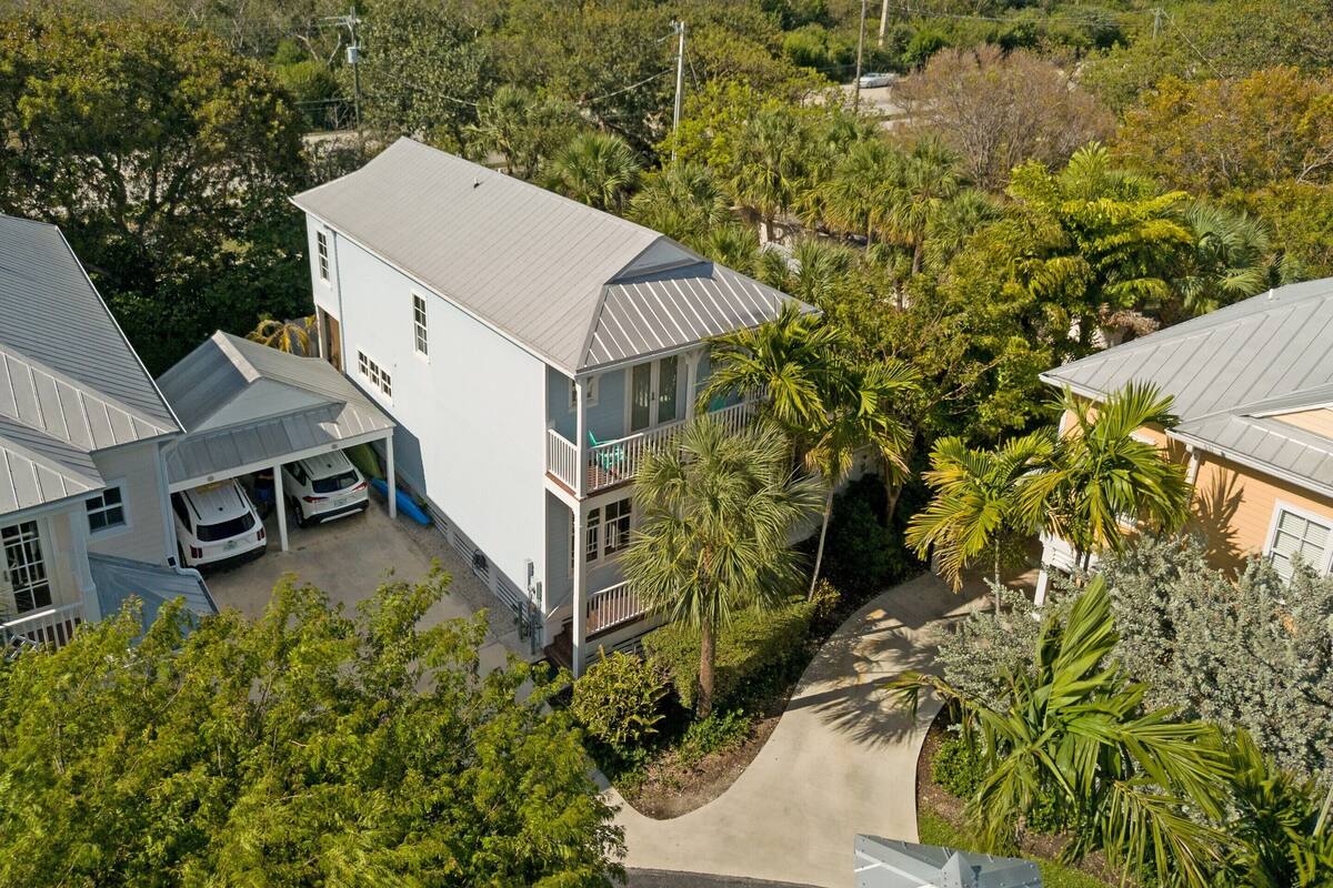 A two-story house with a covered carport and a balcony, surrounded by lush greenery and trees.