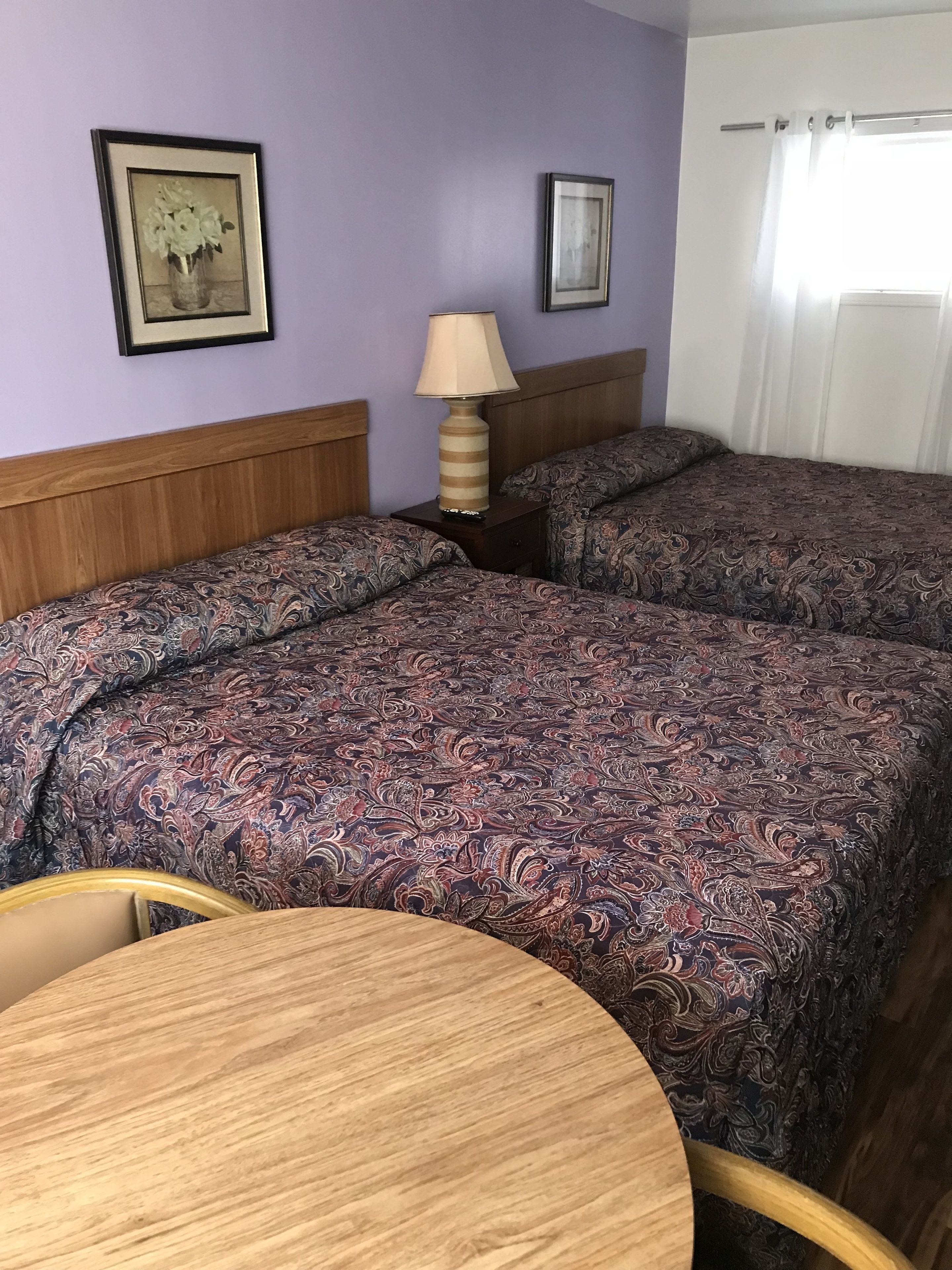 Ein Hotelzimmer mit zwei Betten, einem Holzkopfende, einem Nachttisch mit Lampe, einem gerahmten Bild an der Wand und einem runden Holztisch.