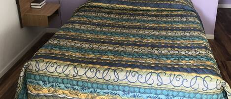 Una cama con una colcha estampada, un cabecero de madera, una mesita de noche con una lámpara y un cuadro enmarcado en la pared.