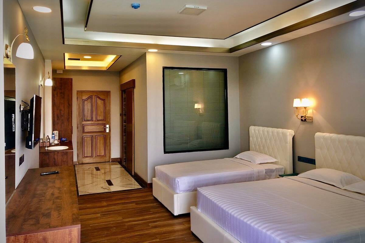 A hotel room with two beds, a wooden desk, a mirror, a television, and a window with blinds.