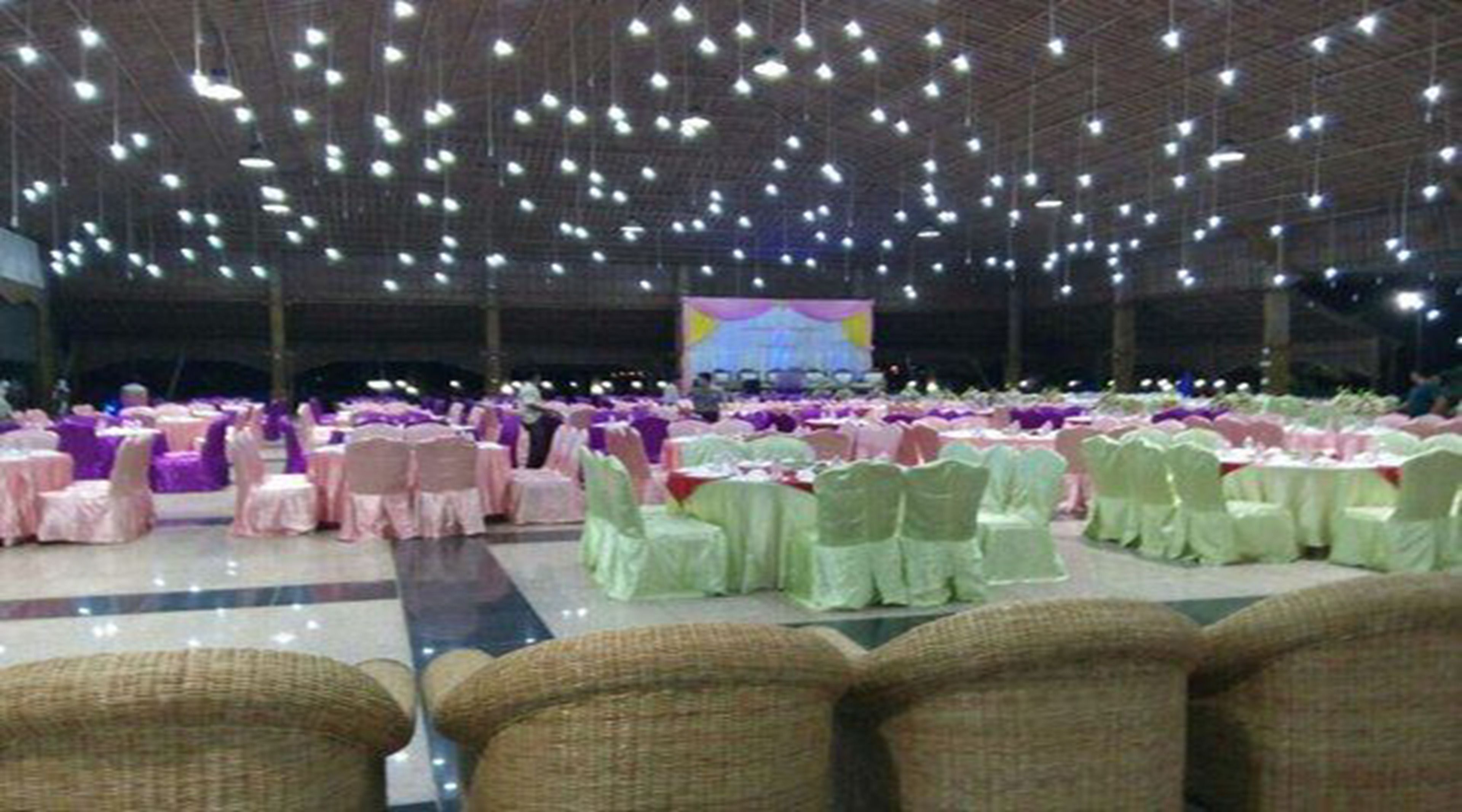 Un amplio espacio interior para eventos, con filas de mesas y sillas, un escenario con un telón de fondo y numerosas luces colgantes.