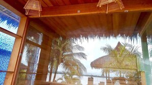 A room with wooden ceiling beams, a large window with a view of palm trees and a thatched-roof structure, and a hanging light fixture. - Excel River View Hotel (Thanlyin)