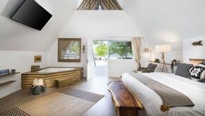 A modern bedroom with a large bed, a wooden bench, a bathtub, and a TV mounted on the wall.