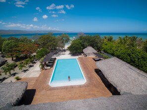 A pool with a wooden deck, surrounded by thatched umbrellas and lounge chairs, overlooking a beach and ocean.