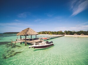 A boat docked at a pier with a thatched-roof structure, clear turquoise water, and a beach in the background.
