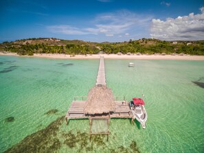 An aerial view of a beach with a wooden pier extending into clear turquoise waters, a thatched-roof structure at the end, and a small boat docked nearby.