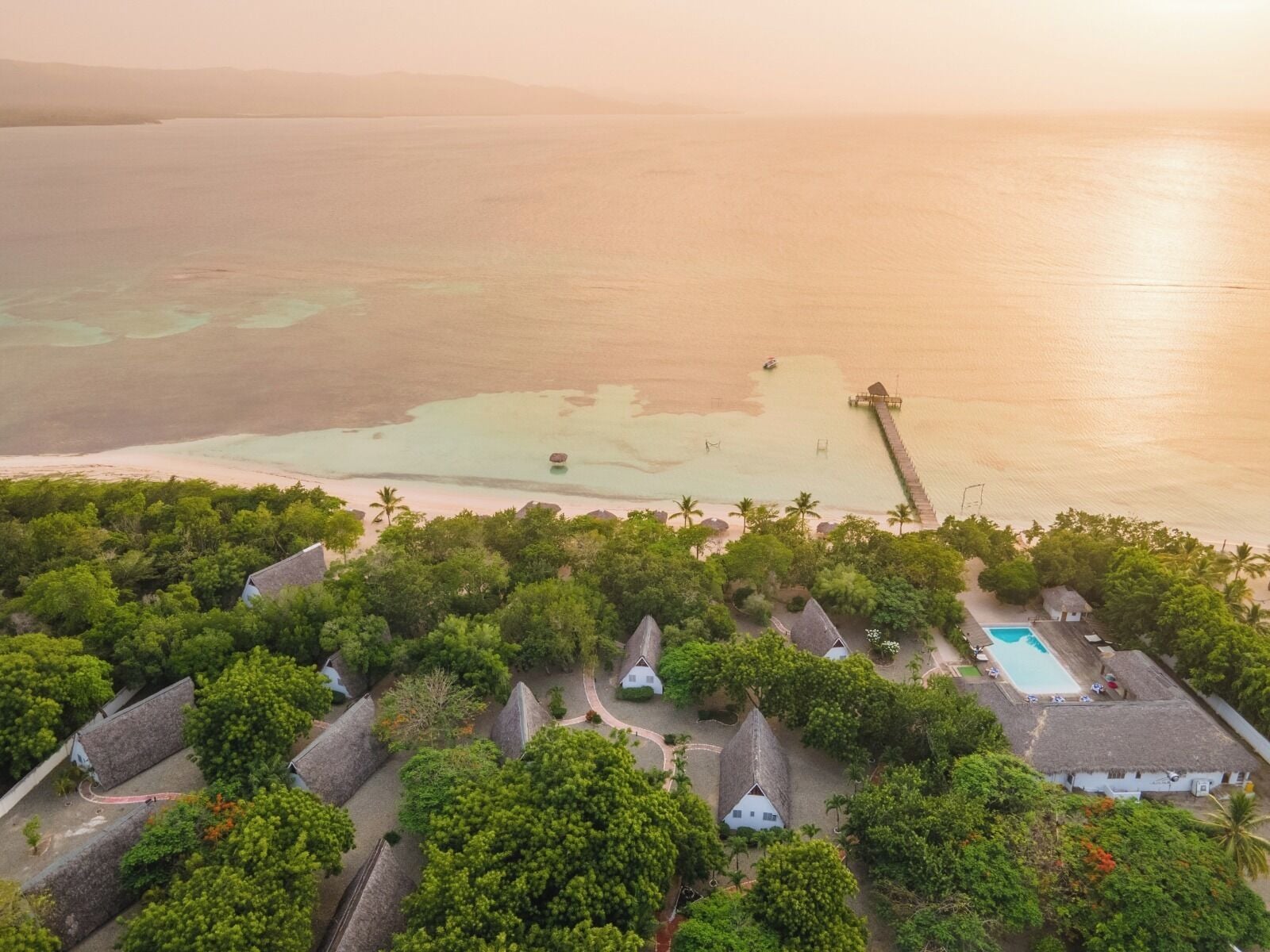 An aerial view of a resort with thatched-roof cottages, a swimming pool, and a pier extending into the sea.