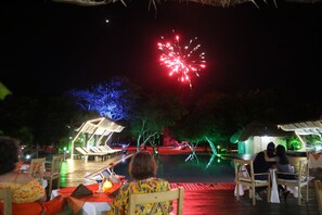 An outdoor dining area with tables and chairs, a thatched-roof structure, and a view of fireworks in the background. - Punta Rucia Lodge Hotel Boutique & Spa (Punta Rucia)