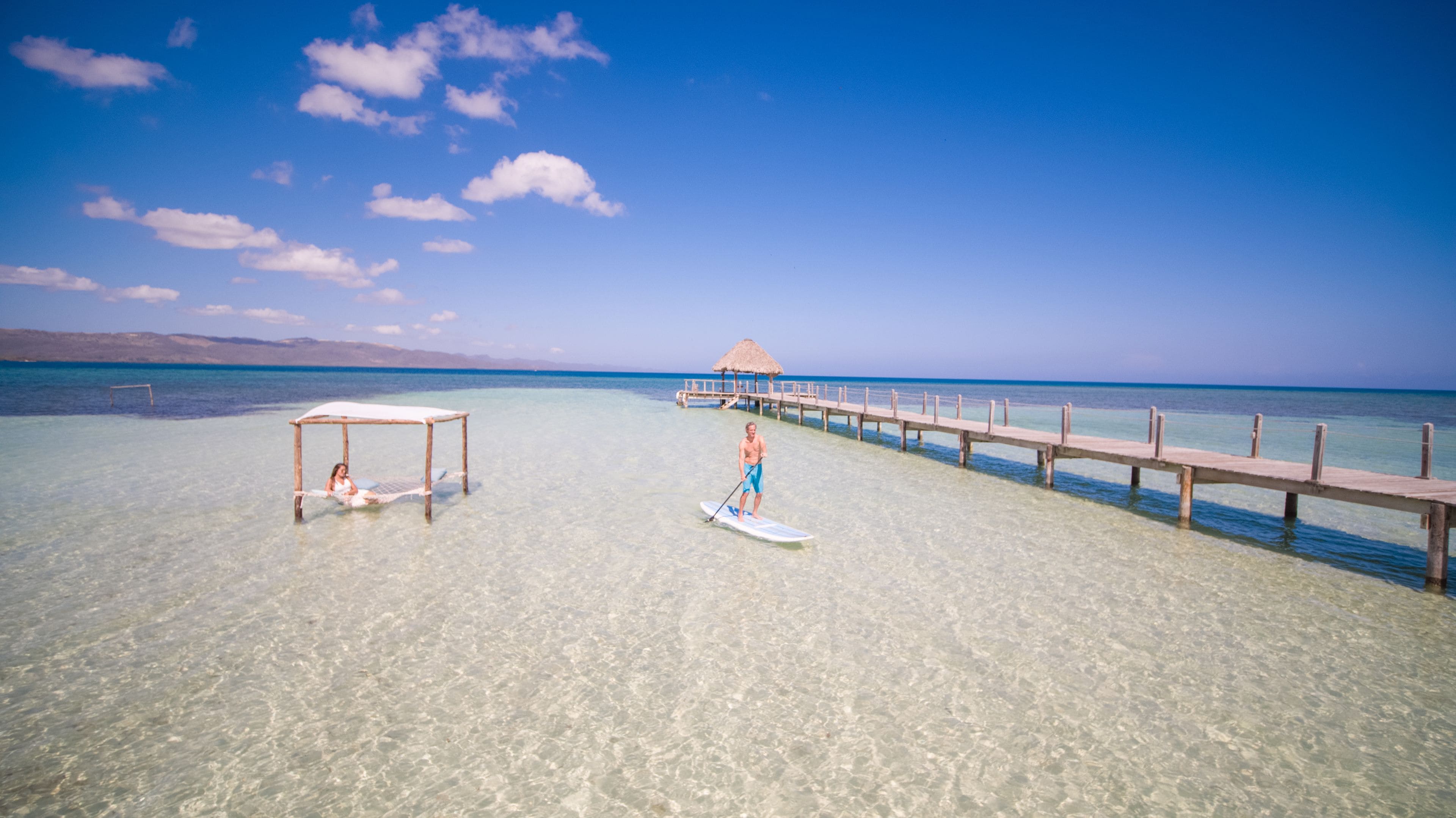 Una playa con agua cristalina, un muelle de madera y una cabaña con techo de paja.