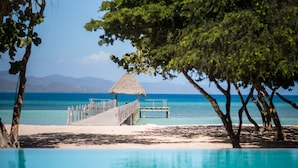 A beach with a wooden pier leading to a thatched-roof structure, surrounded by trees and a clear blue sea.