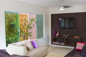 Una sala de estar moderna con un sofá, un televisor montado en la pared y un ventanal con vistas a zonas verdes.