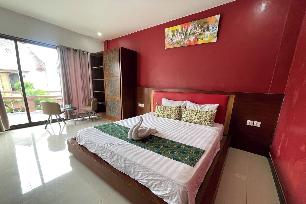 A hotel room with a large bed, a wooden headboard, a painting on the wall, and a view of the outside through a sliding door.