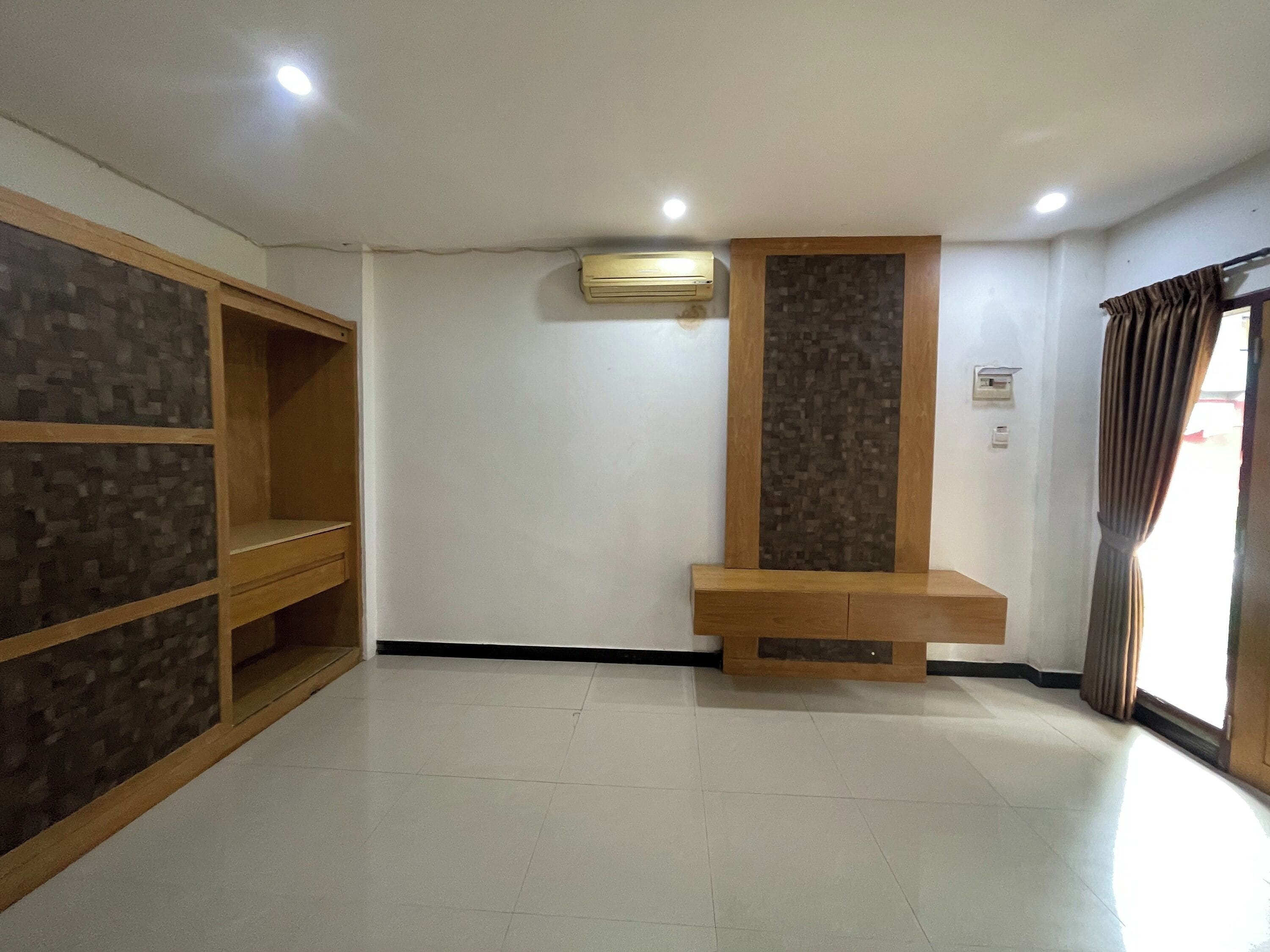A room with a tiled floor, a built-in wooden shelf unit, a wall-mounted air conditioning unit, and a window with curtains.