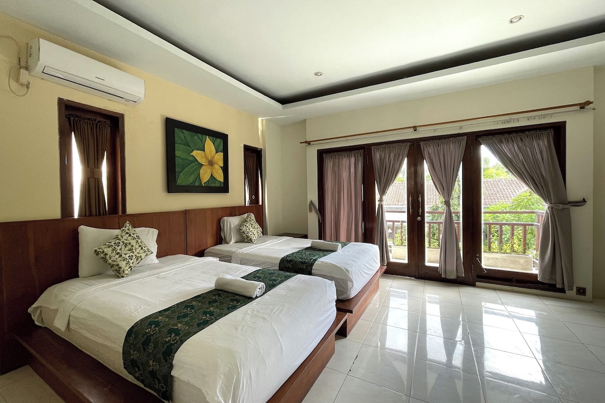 A hotel room with two beds, a large window with curtains, a ceiling fan, and a wall-mounted air conditioner.