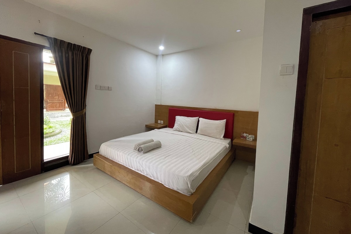 A hotel room with a bed, a nightstand, a wooden headboard, a door, and a window with curtains.