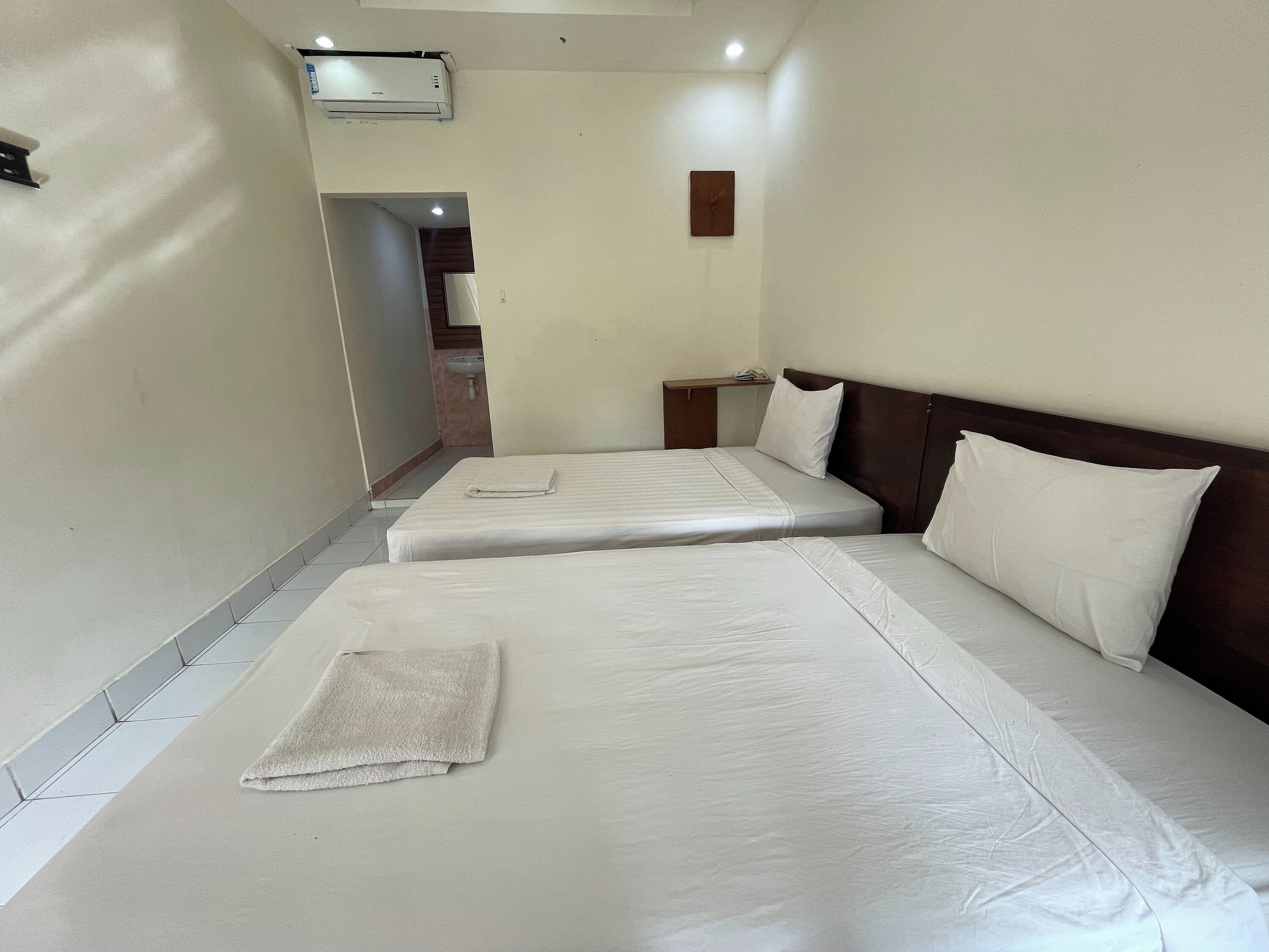 A hotel room with two beds, a bedside table, a wall-mounted clock, and an air conditioning unit.