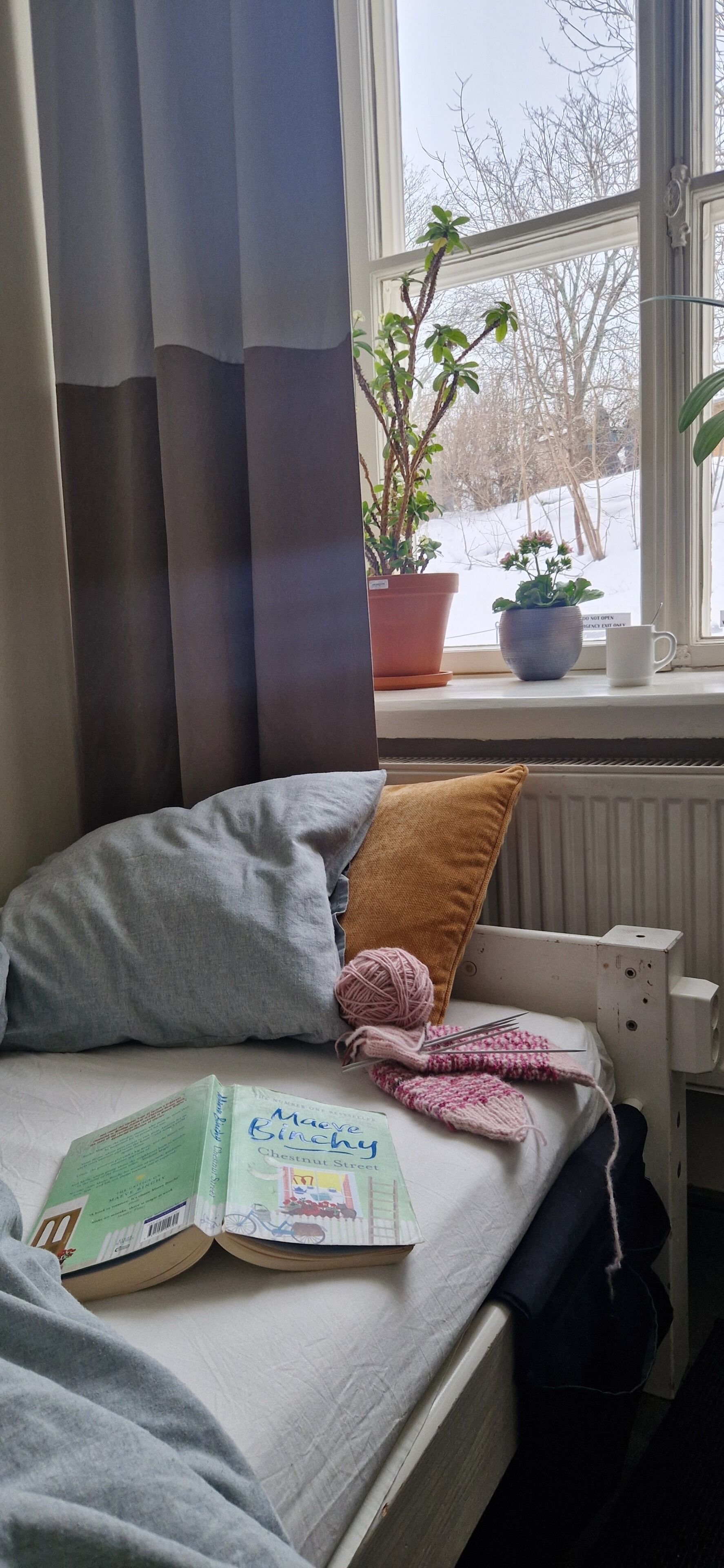 Un dormitorio con cama, ventana con cortinas, radiador, planta y un libro.