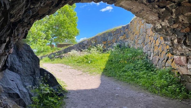 A natural cave entrance with a dirt path leading outside, surrounded by greenery and a stone wall. - Hostel Suomenlinna (Helsinki)