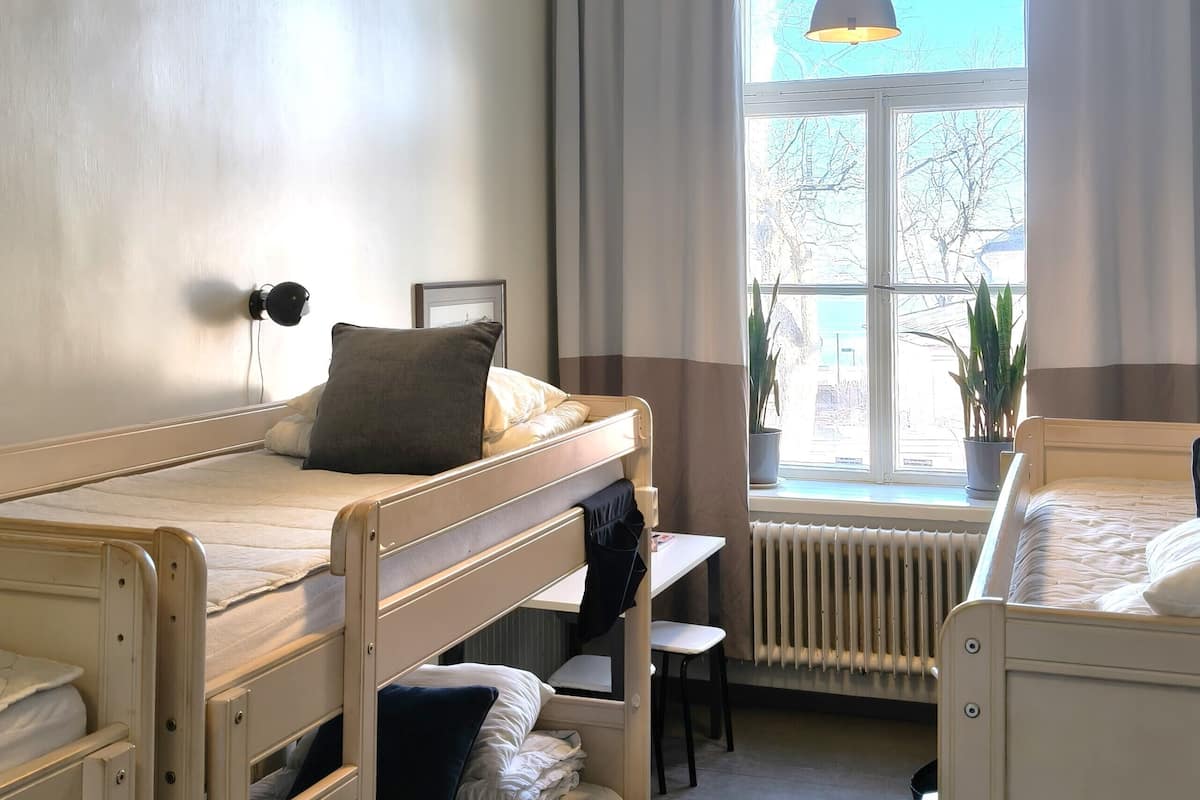 A dorm-style room with bunk beds, a desk, a chair, a hanging lamp, a radiator, and a window with curtains.