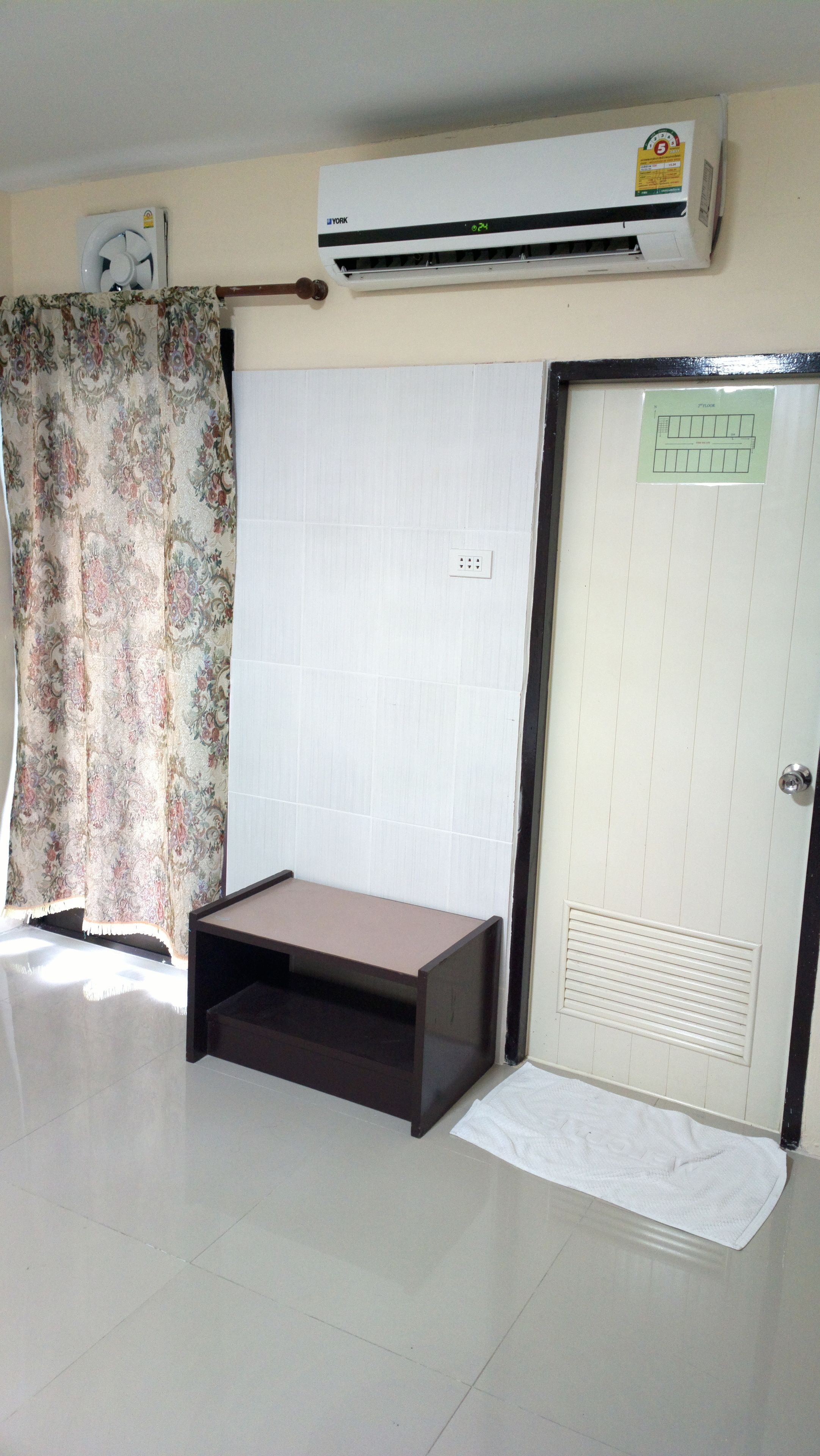 A room with a white air conditioning unit, a door with a floor plan, a patterned curtain, and a small wooden table.